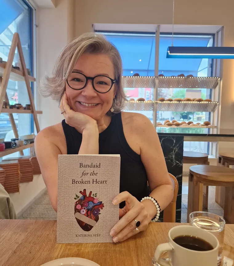 Katerina Szép smiling at a café table, holding a copy of Bandaid for the Broken Heart